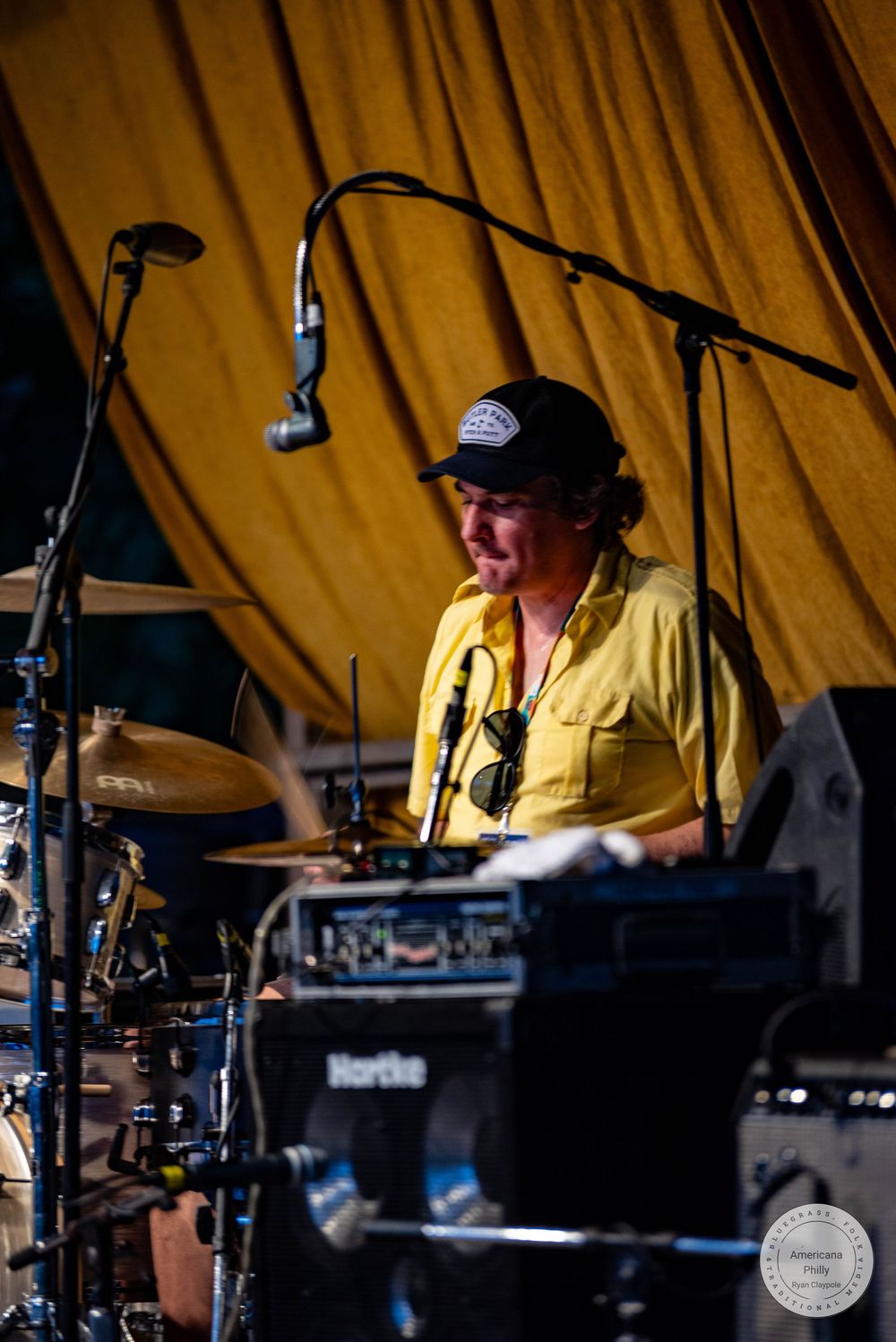 Drummer in yellow shirt and black cap performing on stage with cymbals and microphones at an outdoor music event.