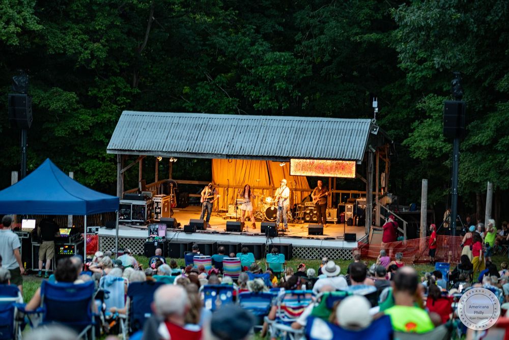 Outdoor music festival with a rustic wooden stage, live band performing, and a large crowd seated on a lawn surrounded by trees.