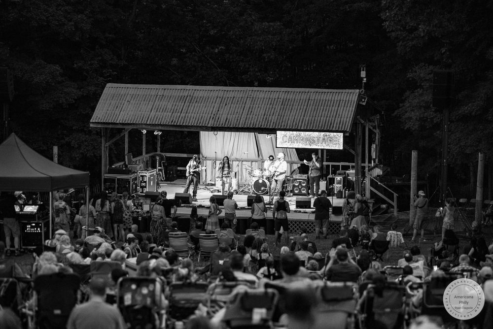 Outdoor music festival with a band performing on a rustic wooden stage surrounded by trees and a large crowd.