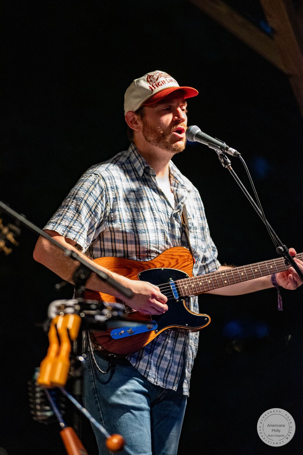 Man in plaid shirt and cap playing guitar and singing into microphone on stage at outdoor concert.