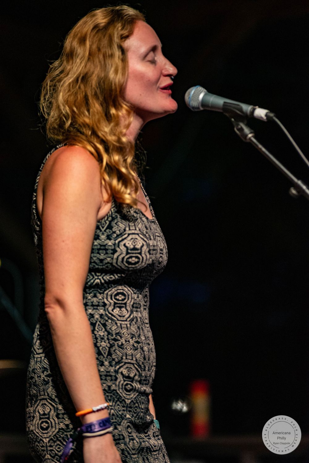 Woman with curly hair singing into a microphone on stage, wearing a patterned sleeveless dress and colorful bracelets.