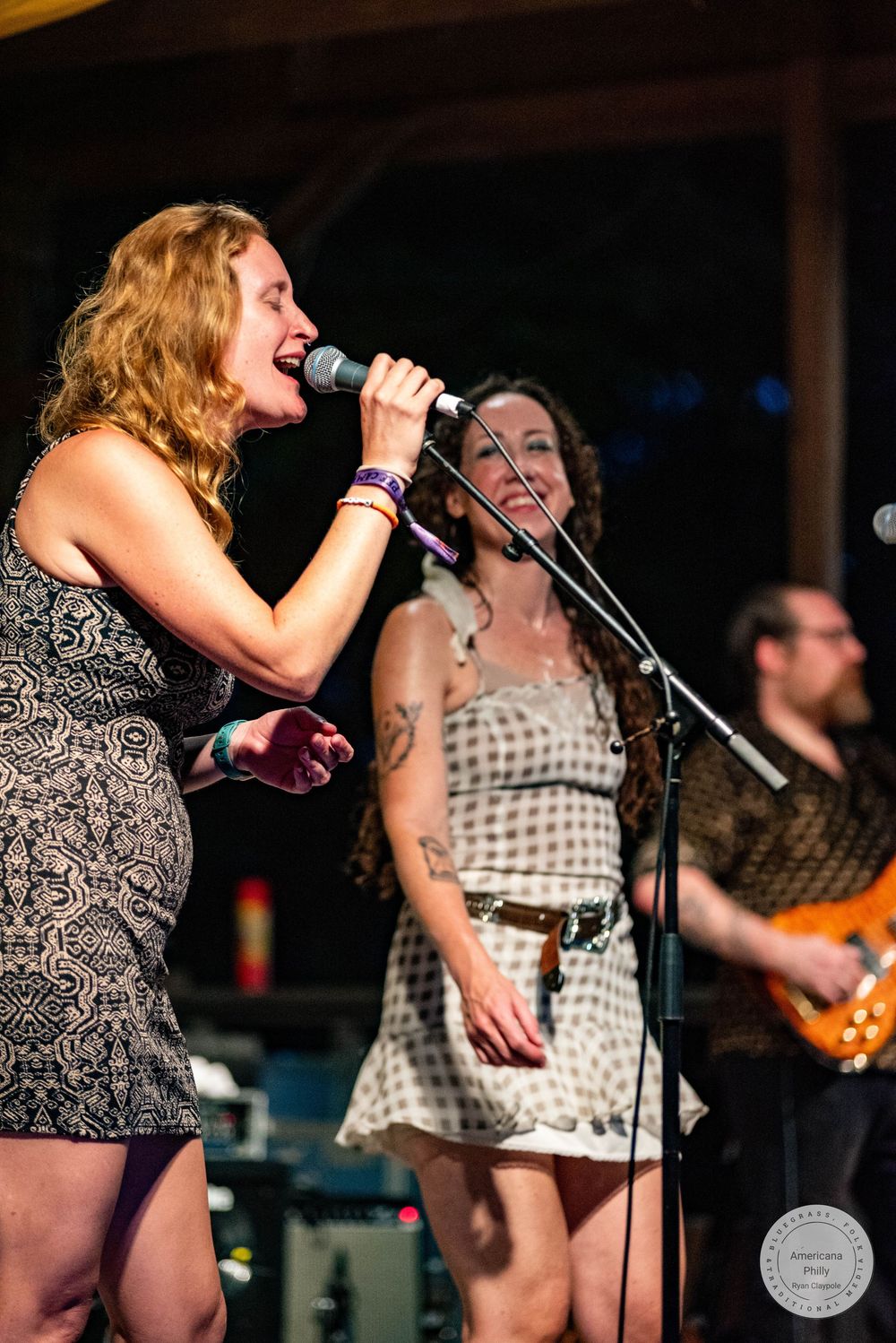 Woman singer belting into microphone on stage with female backup singer and guitarist performing live.