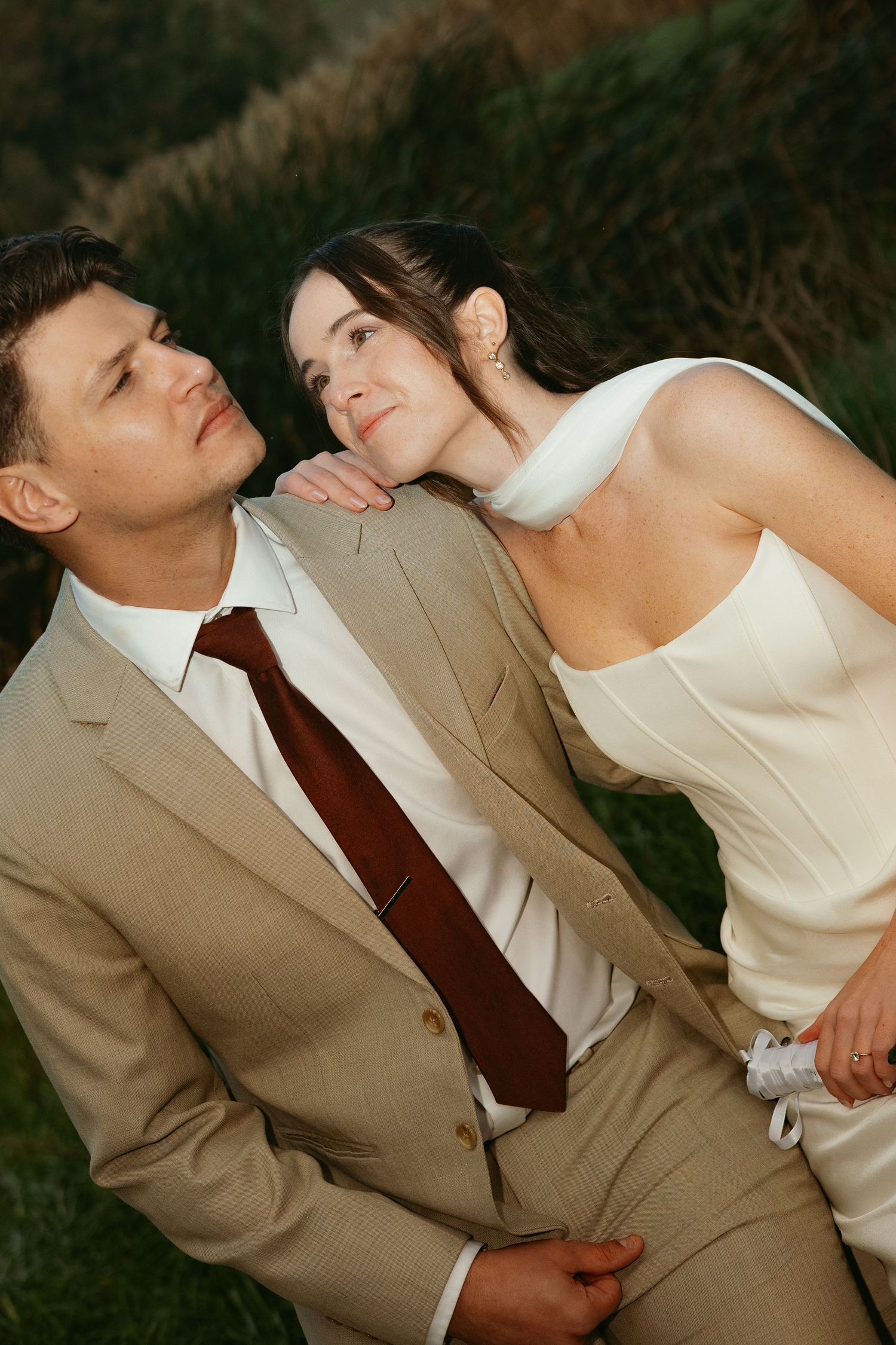 A couple in wedding attire, groom in tan suit with burgundy tie, bride in white dress, posing outdoors near greenery.