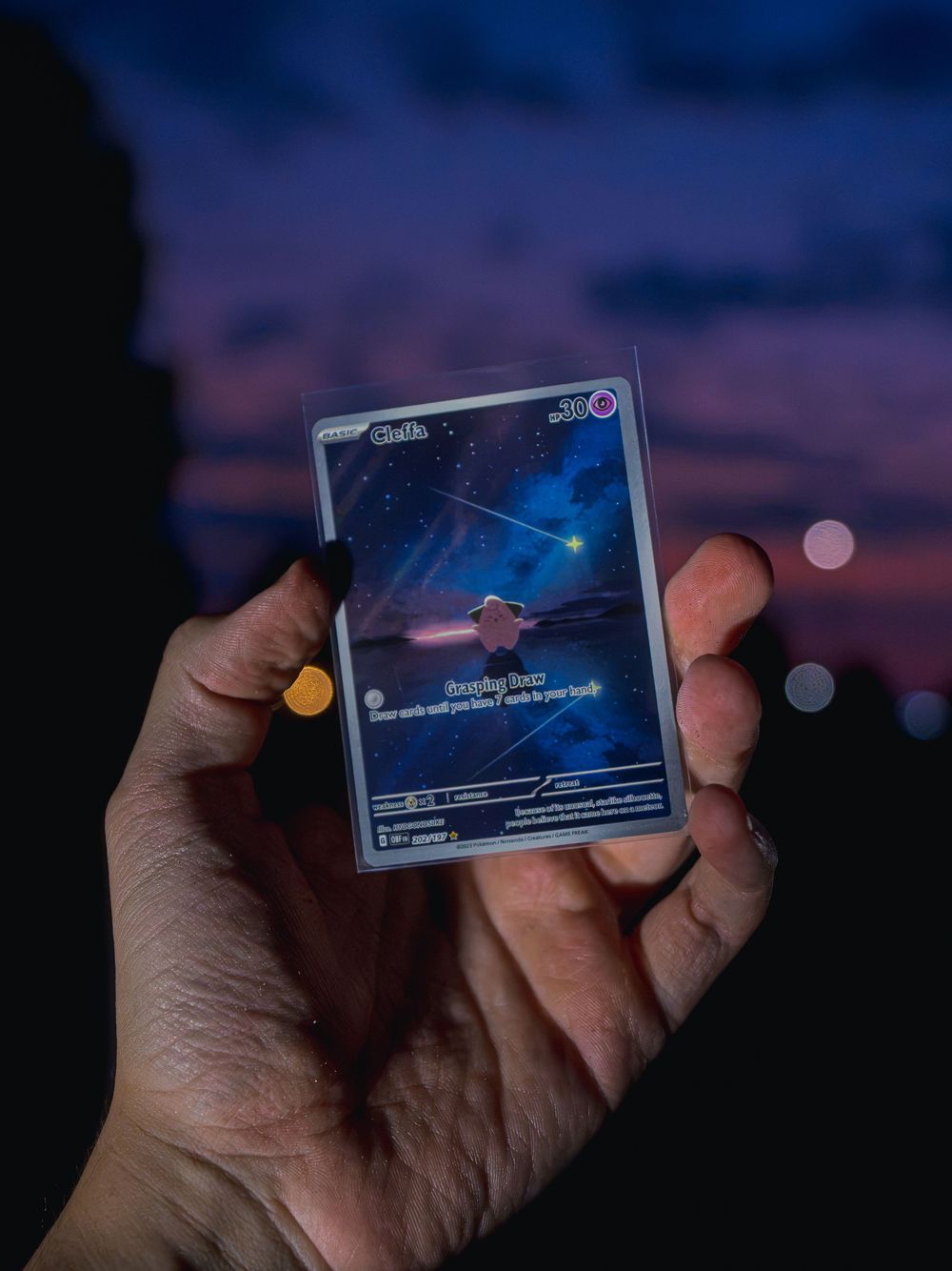 Product photograph of a hand holding up a starry night-sky collectible card against a purple-and-pink dusk sky with soft bokeh lights behind
