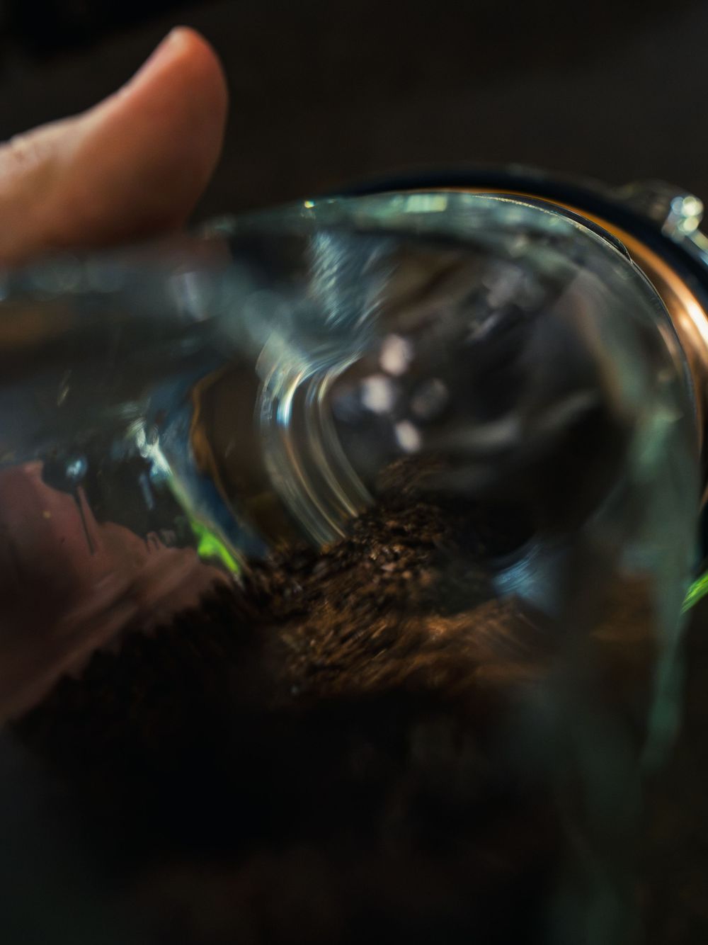A hand tilting a glass of ground coffee, the grounds sliding toward a metal pour-over filter in moody, low light.