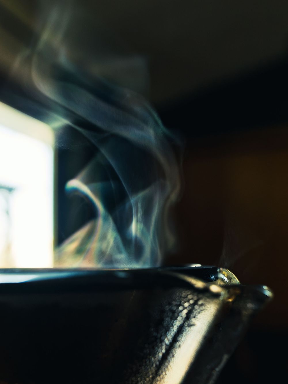 Wisps of steam curling off the rim of freshly brewed coffee, backlit by soft window light against a dark background.