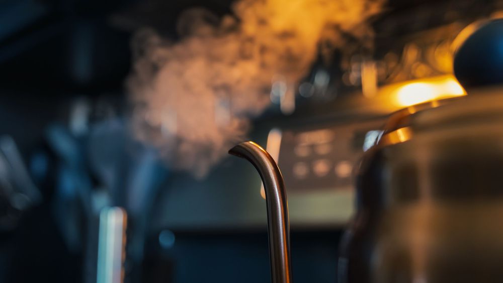 Steam rising from a gooseneck kettle spout, water heated and ready for a pour-over coffee, warm light glowing behind.