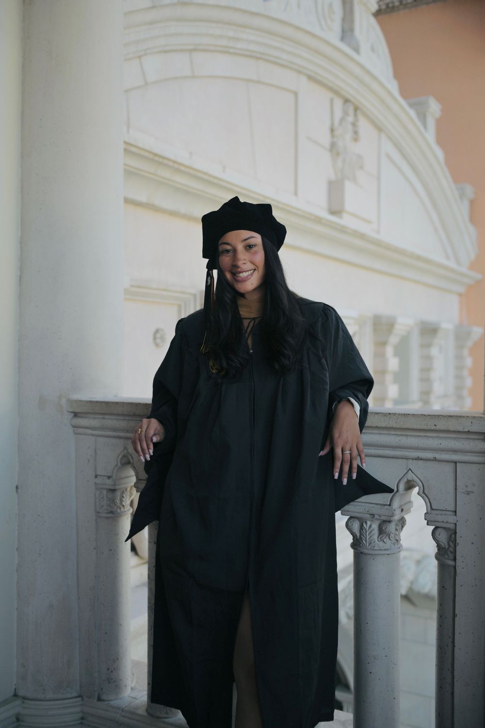 Smiling woman in black graduation gown and cap posing on ornate white stone balcony.