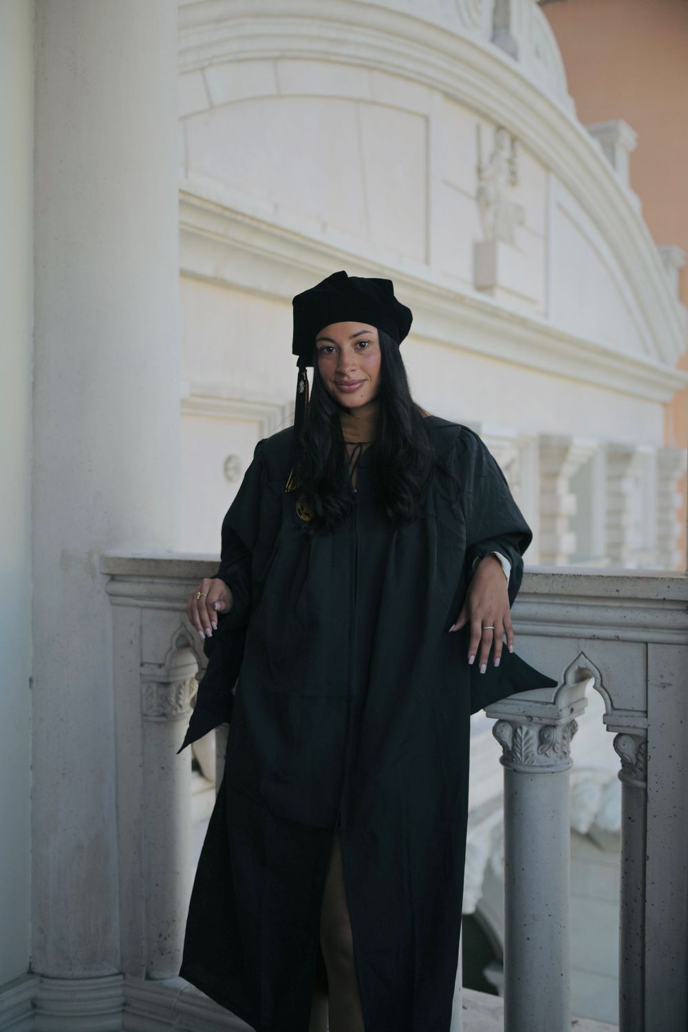 A woman in black graduation gown and cap poses proudly on elegant white stone steps at her graduation ceremony.