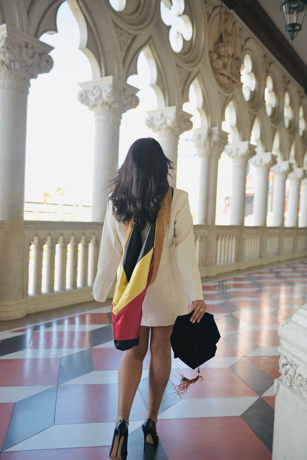 Woman in white blazer with colorful sash walks through ornate marble colonnade with decorative arches.