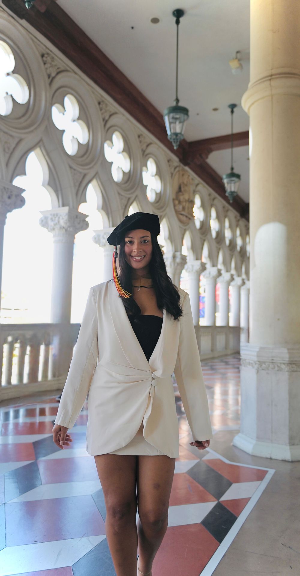 Woman in cream blazer dress and black beret poses in ornate columned hallway with Gothic arches.