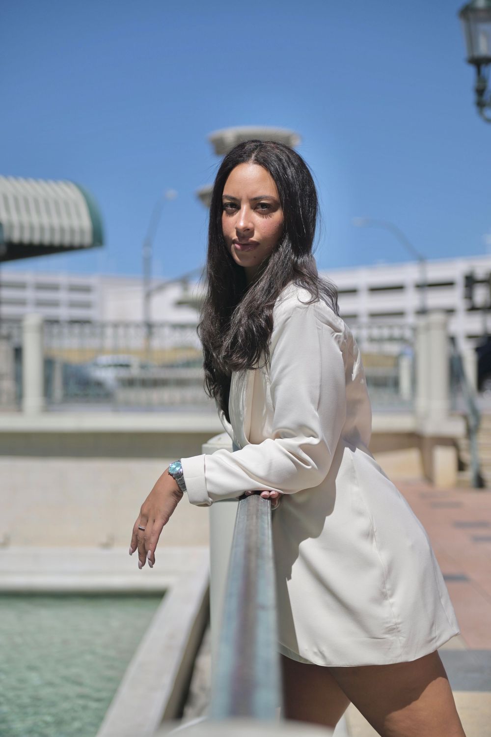 Young woman in white blazer dress leaning on railing outdoors on sunny day.