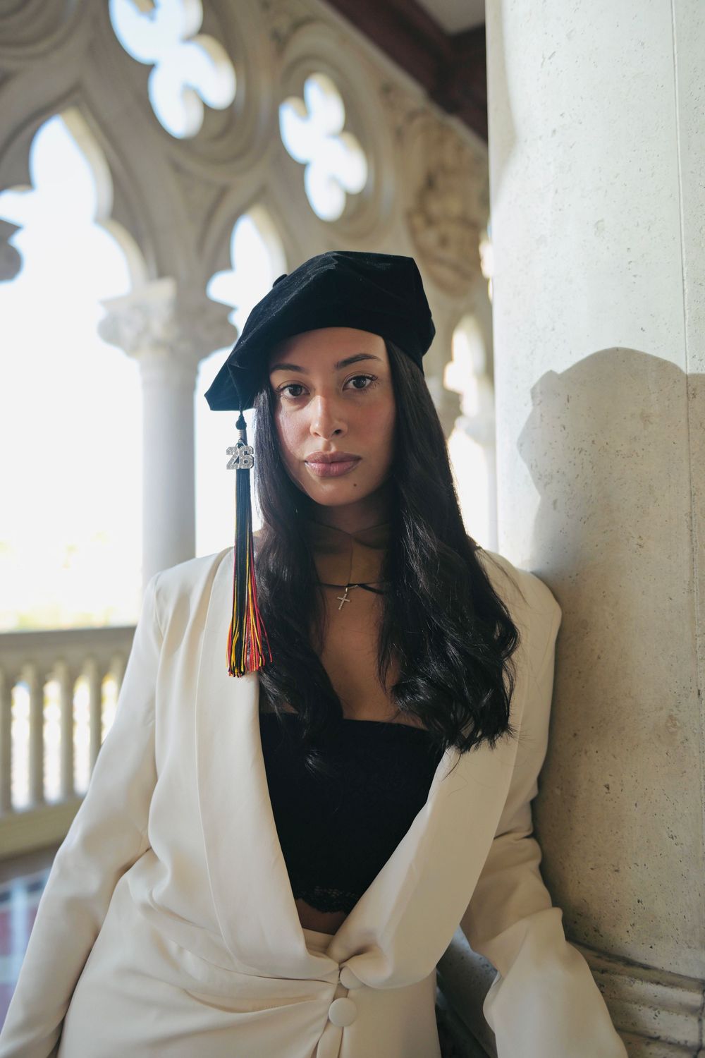 Young woman in graduation cap with colorful tassel and white blazer posing by ornate stone columns.