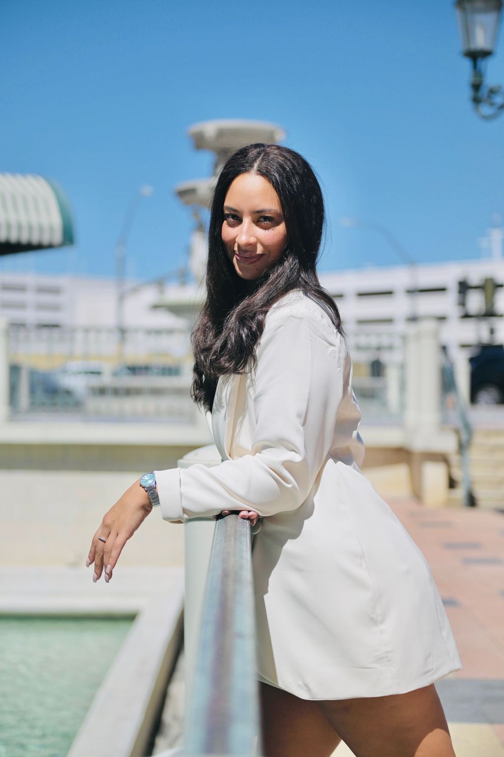Young woman in white blazer dress leans on railing by fountain, smiling outdoors on sunny day.