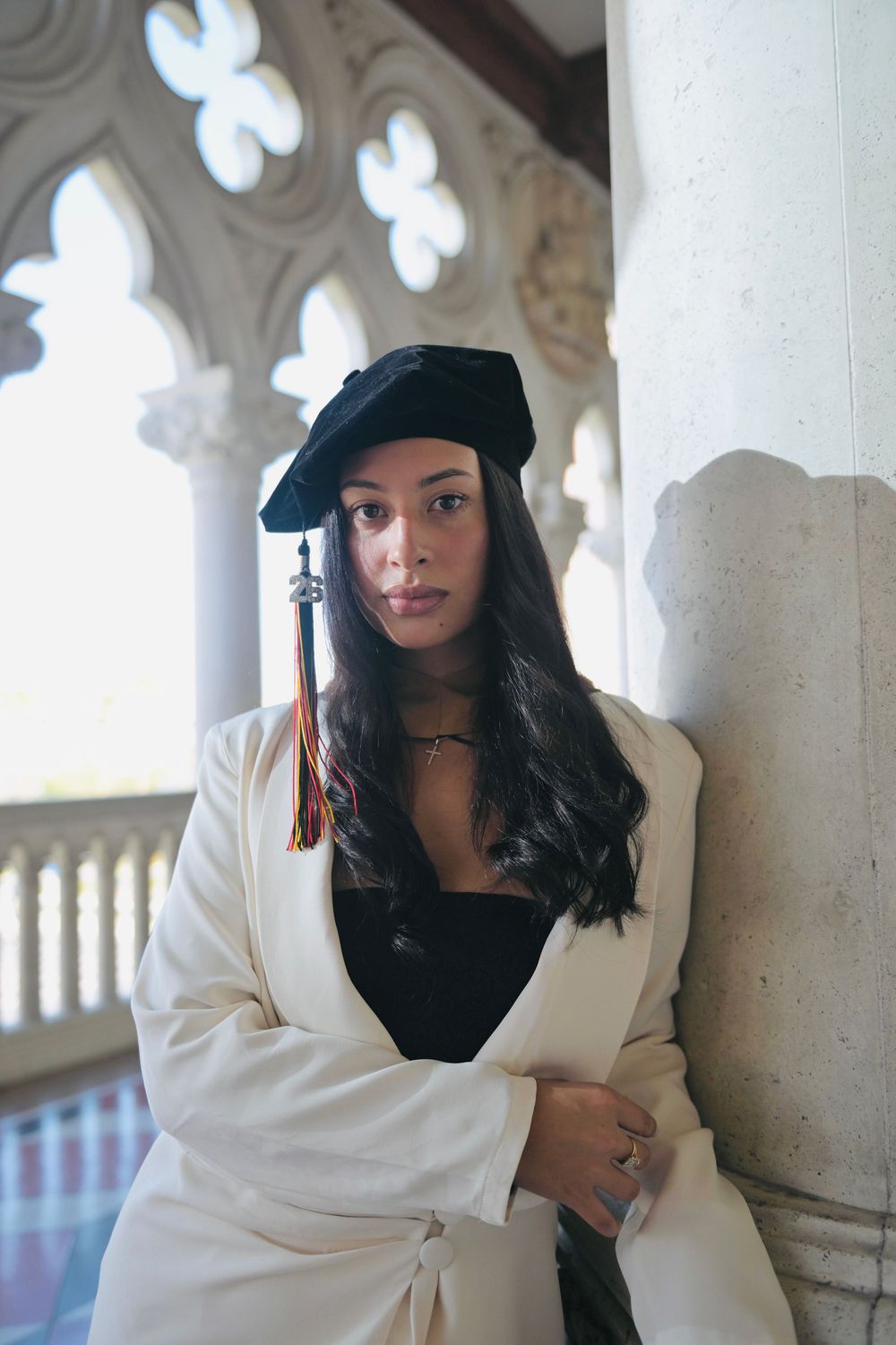 Woman in graduation cap and white robe leaning against stone column indoors.