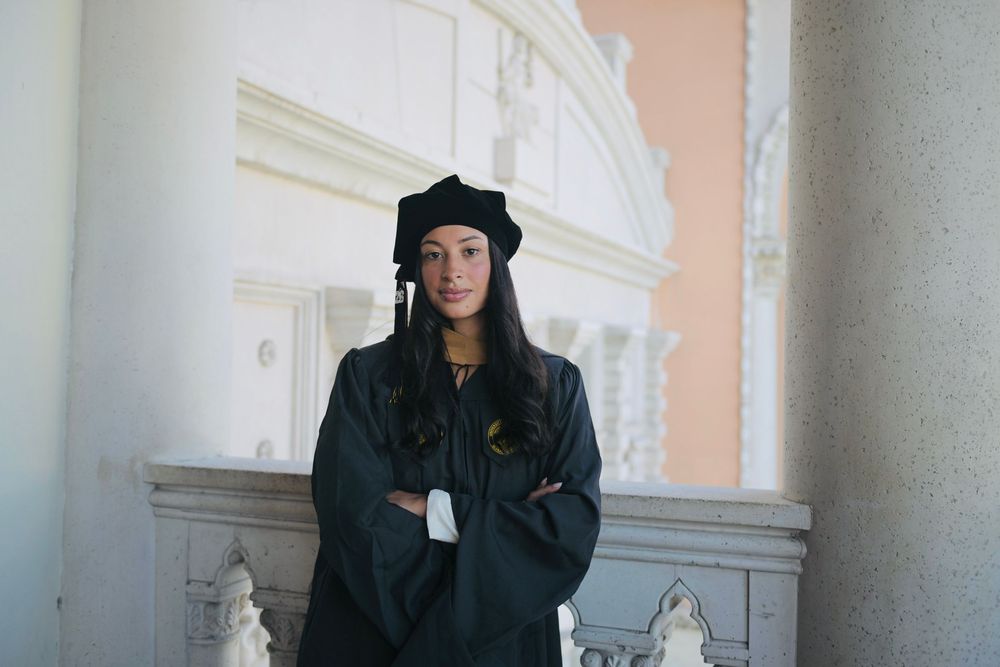 Young woman in black graduation cap and gown stands confidently with arms crossed at a classical building.