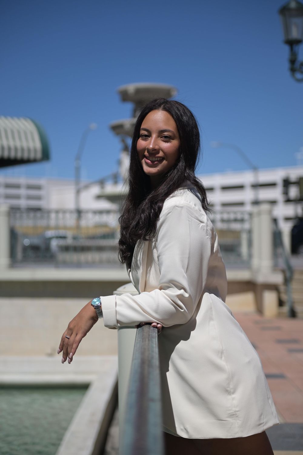 Smiling young woman in white blazer leaning on railing outdoors on sunny day with fountain in background.