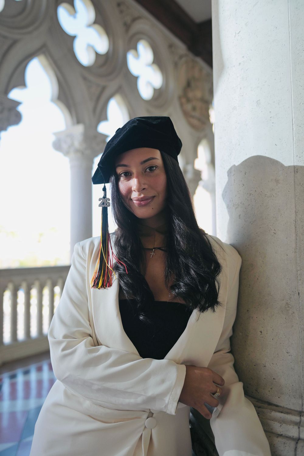 Smiling graduate in black cap and white robe posing by ornate stone pillar.