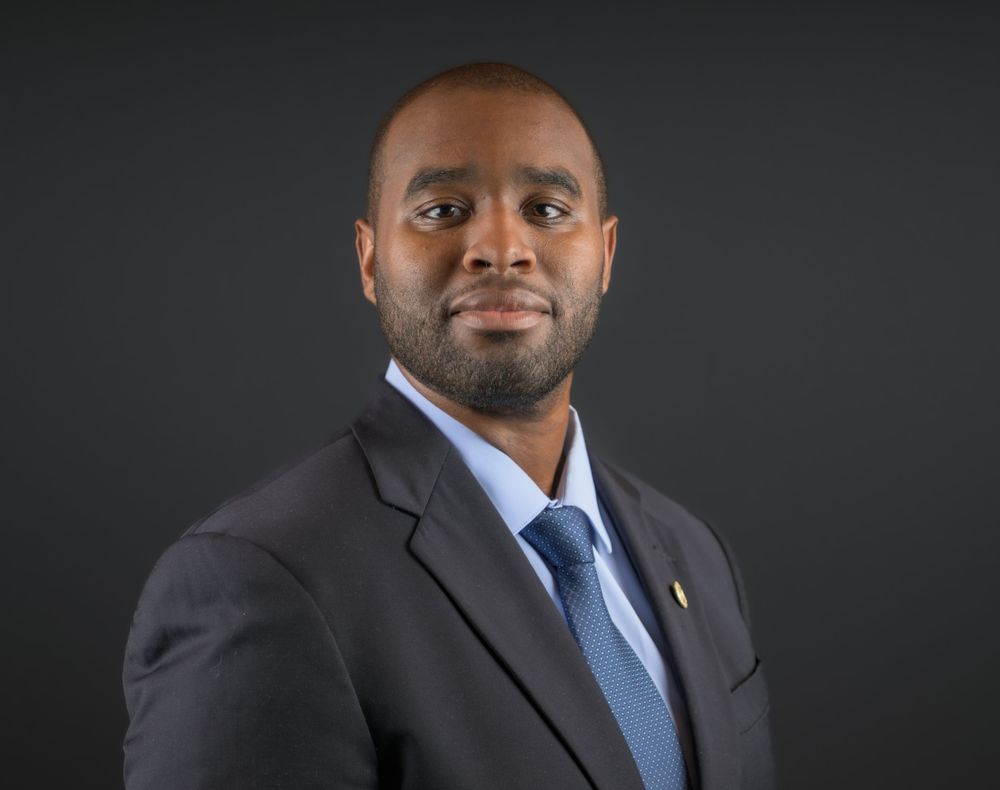 Professional headshot of a man in a dark suit and light blue tie against a gray background.