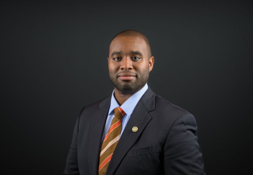 Professional headshot of a man in a dark suit and striped tie against a dark background.