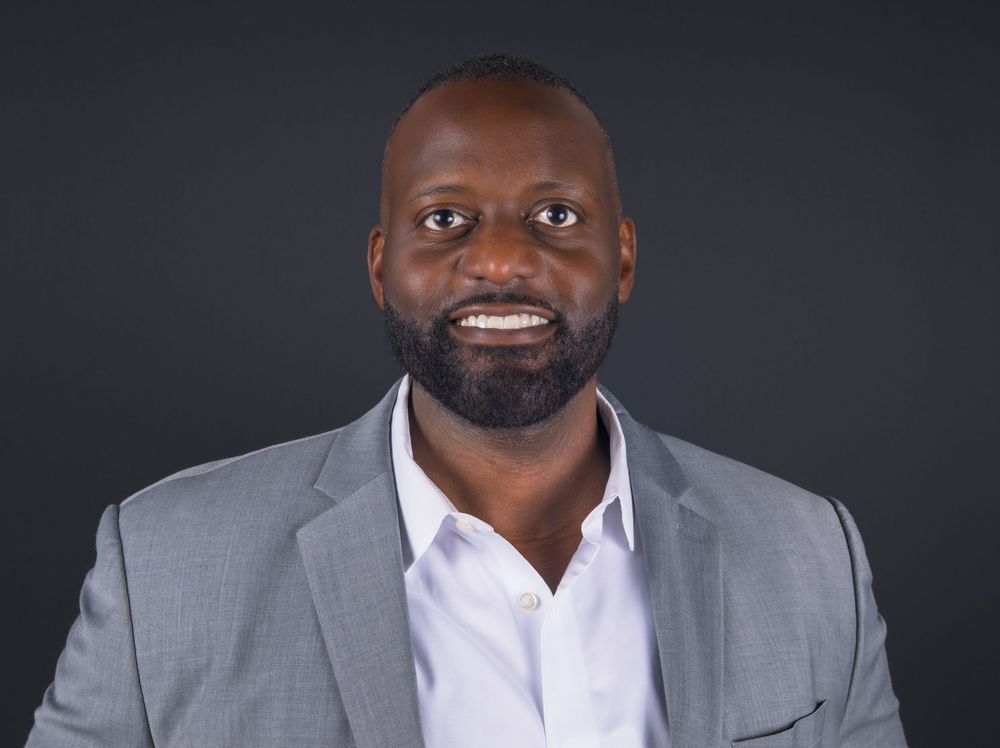 Professional headshot of a smiling man in a gray blazer and white shirt against a dark background.