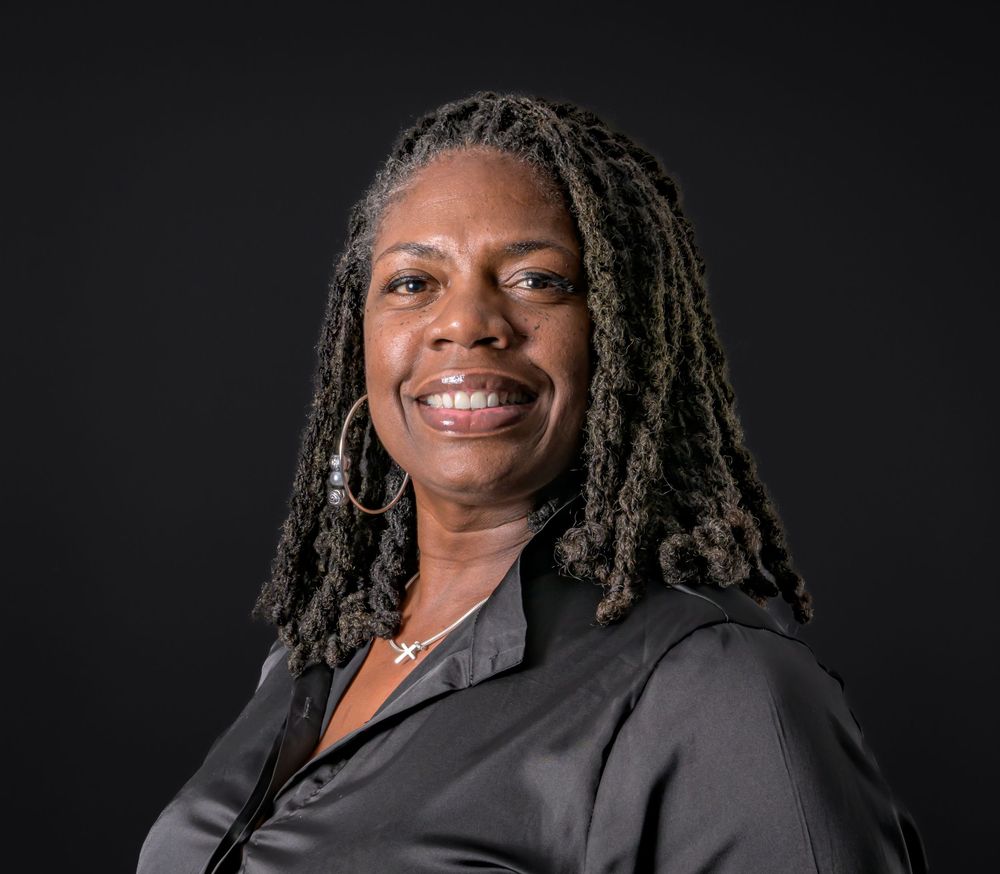 Professional portrait of a smiling woman with locs wearing a black top and hoop earrings against dark background.