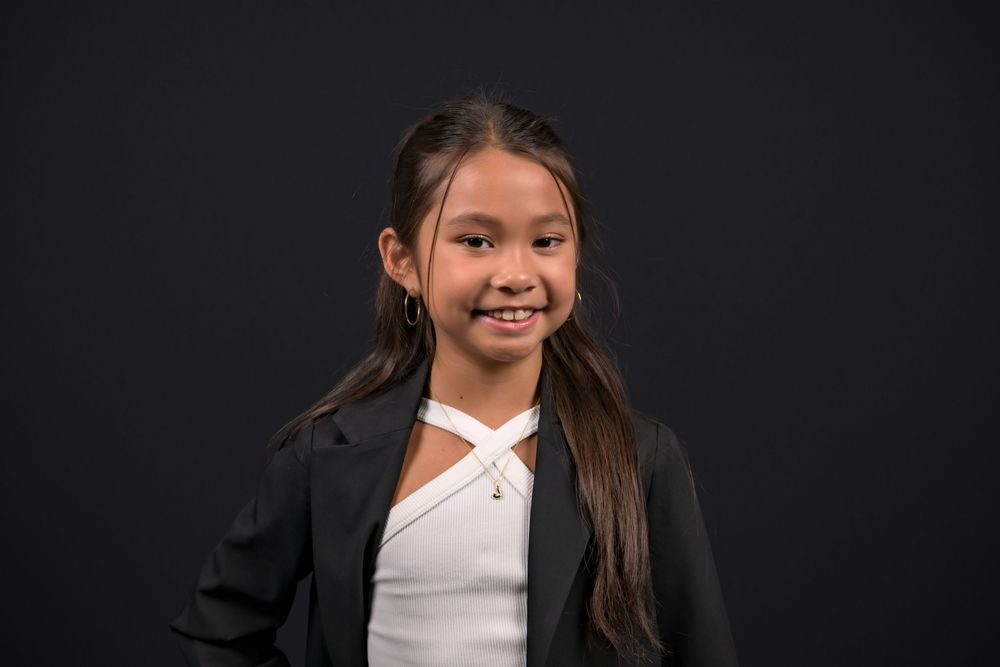 Young girl in black blazer and white top smiling confidently against dark background.