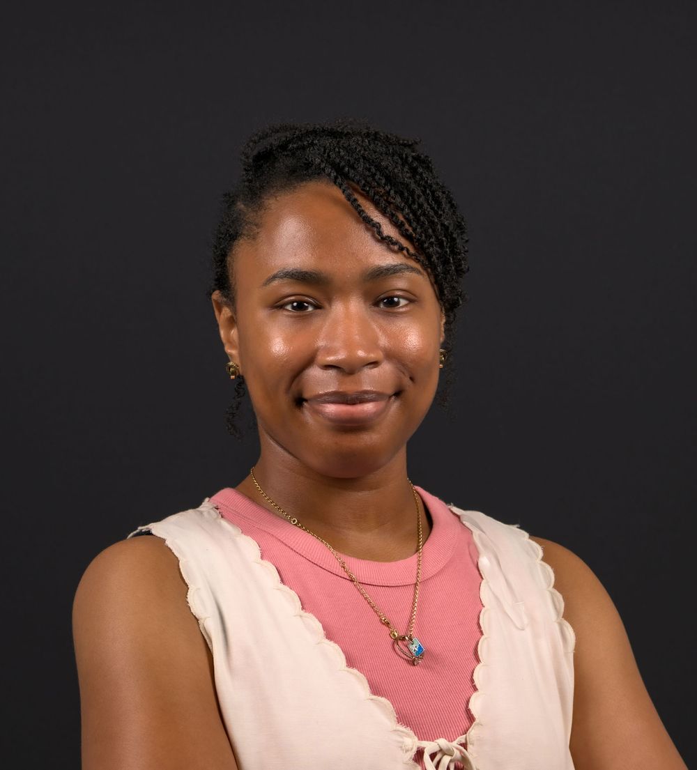 Professional headshot of a young woman with braided hair, wearing a pink top and layered necklaces, smiling warmly.