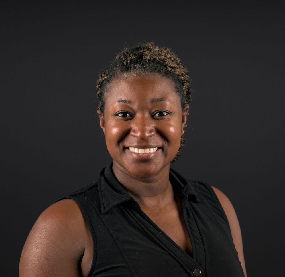 Professional headshot of a smiling woman with short curly hair wearing a black sleeveless top against a dark background.