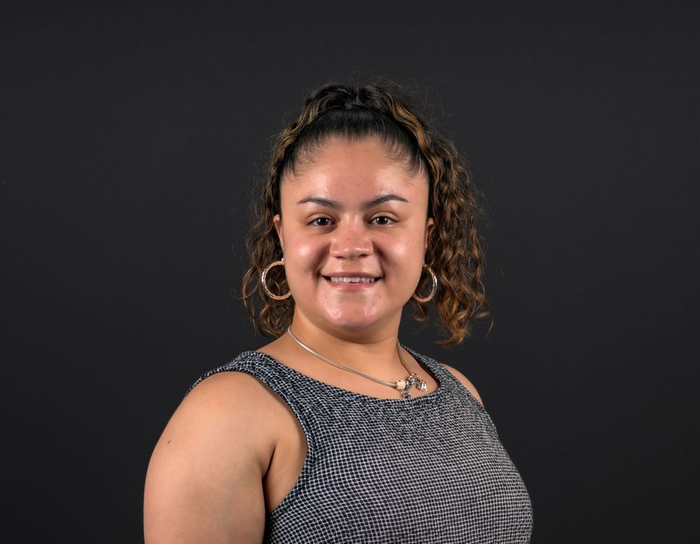 Smiling young woman with curly hair wearing hoop earrings and a gray sleeveless top against dark background.