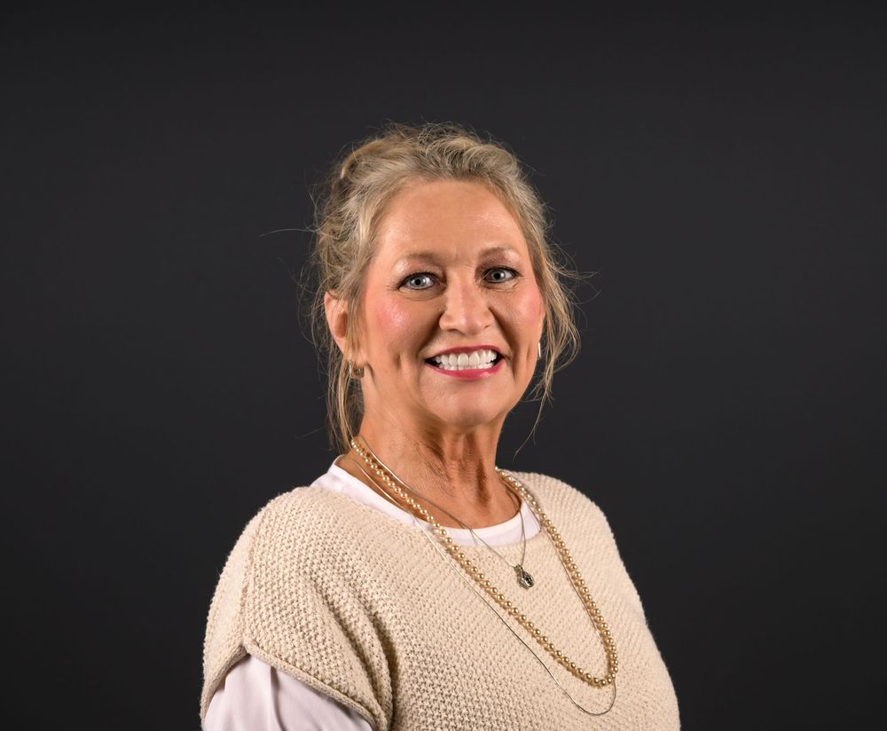 Smiling blonde woman in cream knit vest and layered necklace poses against dark background.