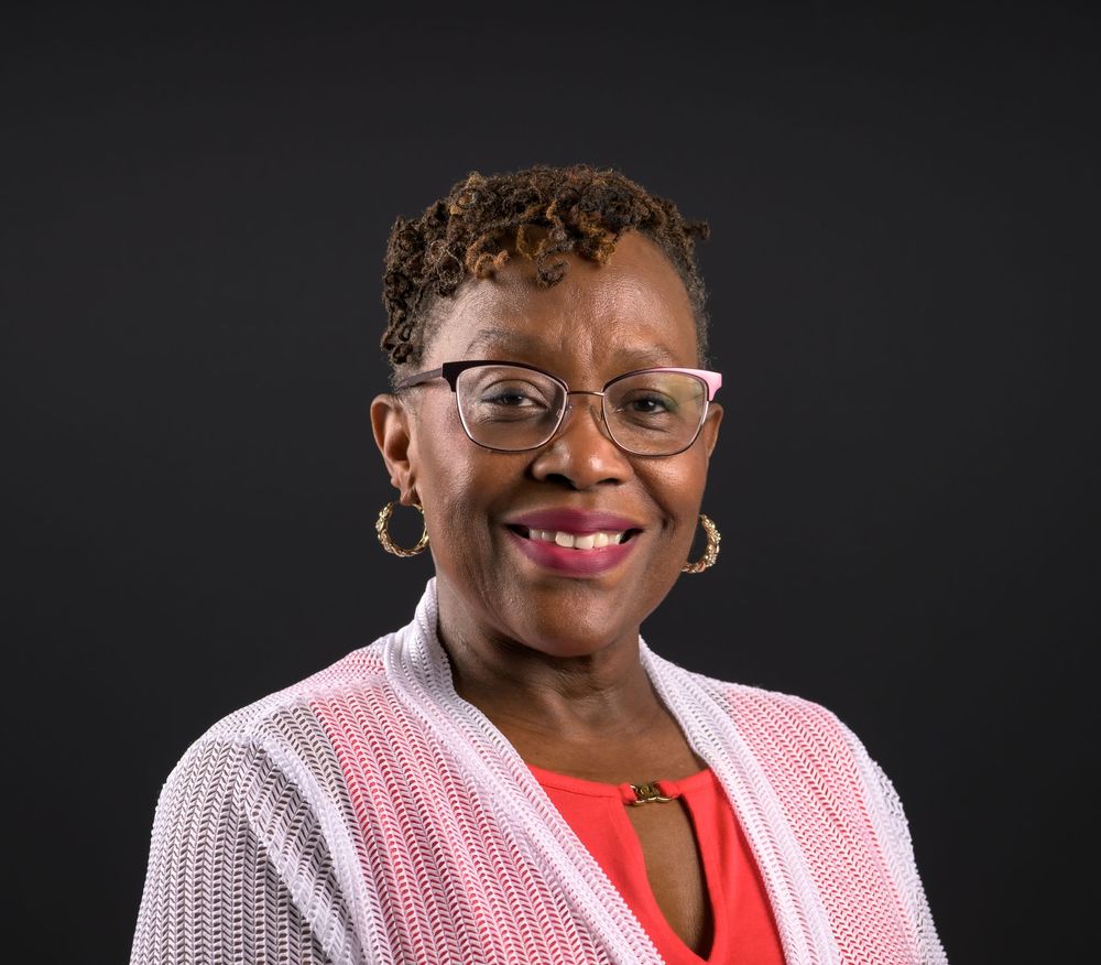 Smiling woman wearing pink glasses, hoop earrings, and a pink cardigan over a red top, posed against a dark background.