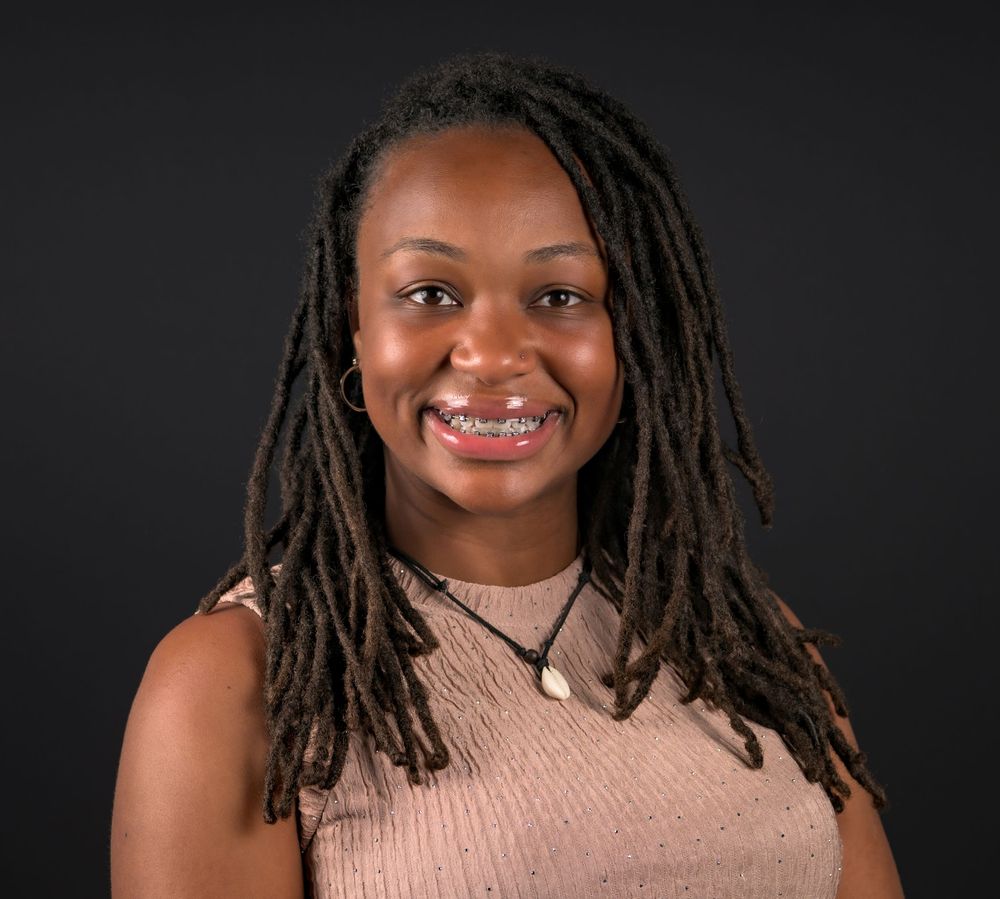 Young woman with locs, wearing a beige top and necklace, smiling confidently against a dark background.