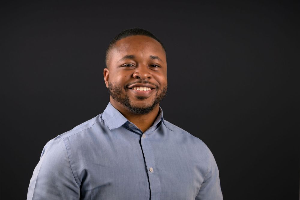 Smiling young Black man in a gray button-up shirt posing against a dark background.