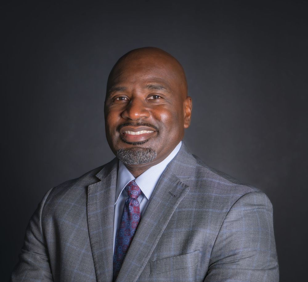 Professional headshot of a smiling man in a gray suit and patterned tie against a dark background.
