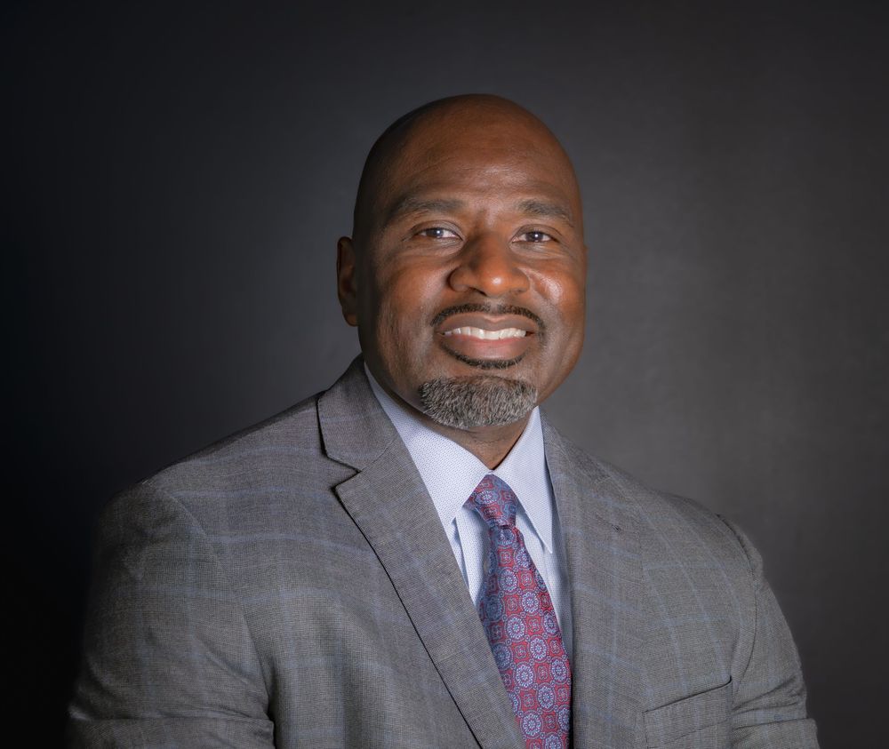 Professional headshot of a smiling man in a gray suit and patterned tie against a dark background.