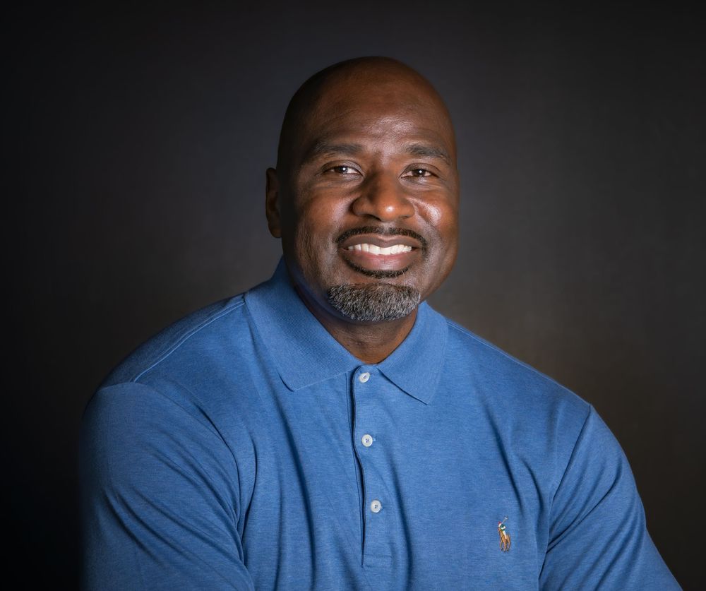 Smiling man in blue polo shirt poses for professional headshot against dark background.