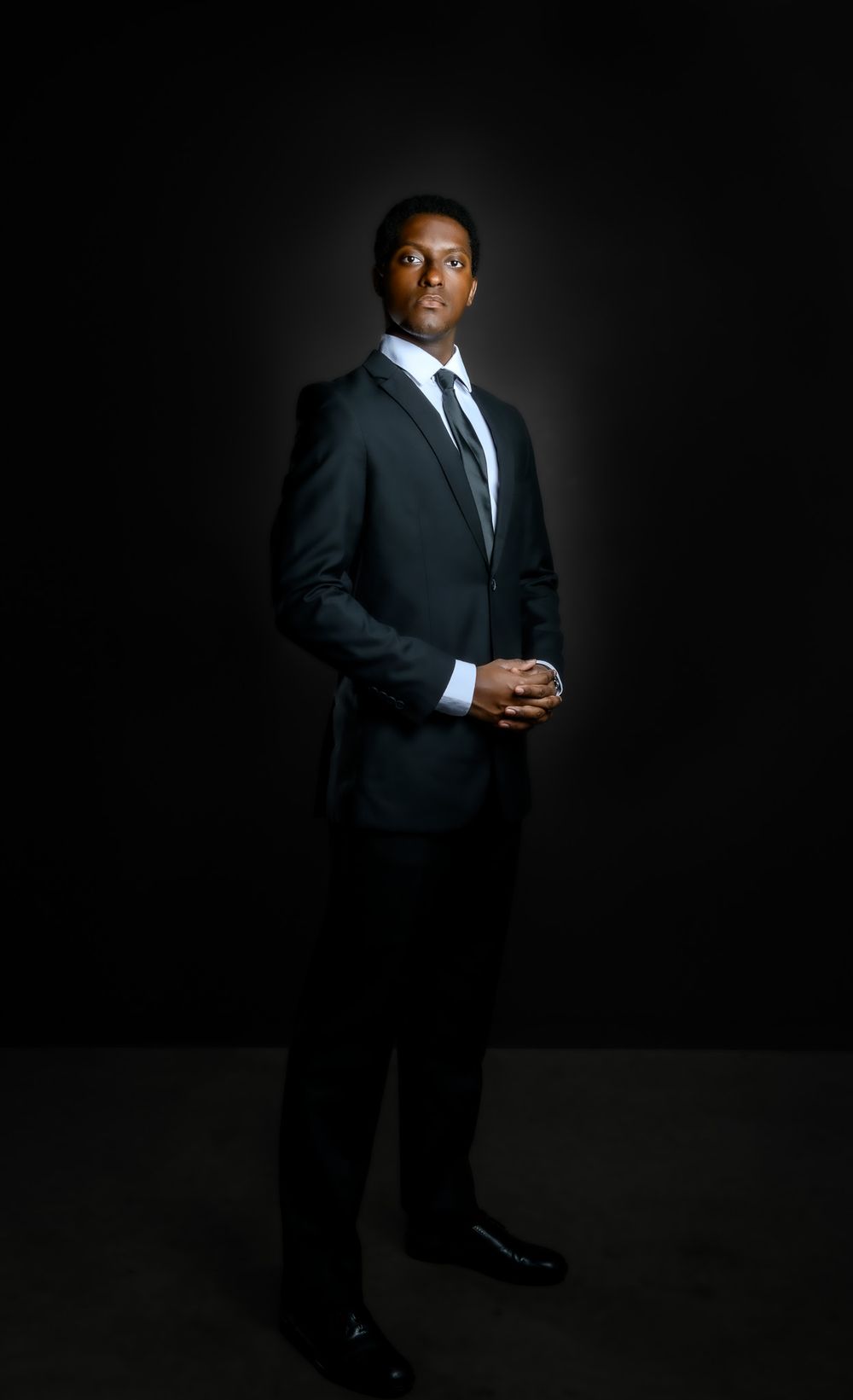 Professional man in dark suit and tie standing confidently against black background in formal portrait.