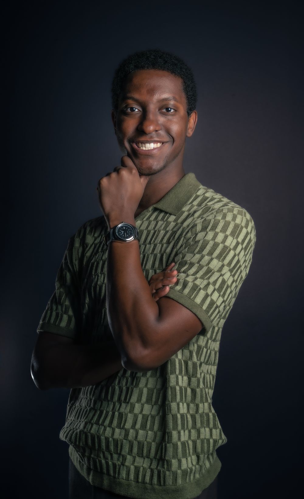 Young man in green patterned polo shirt smiling confidently with arms crossed against dark background.