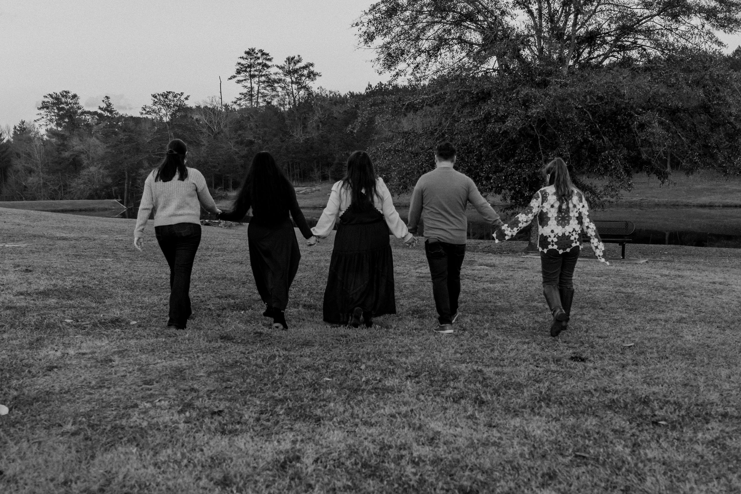Black and white photo of five people holding hands walking across a grassy field toward trees.