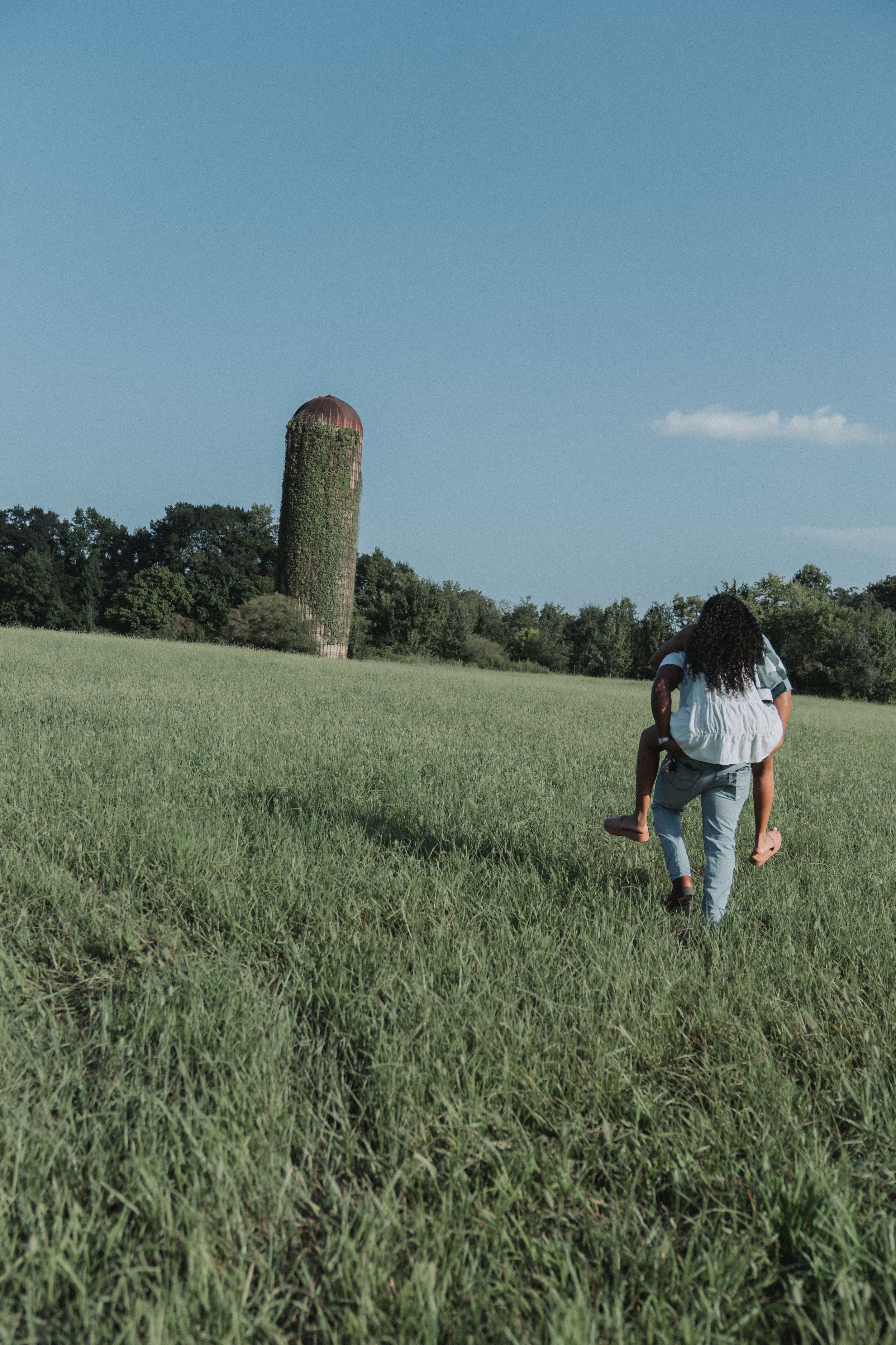 Person carrying someone on their back walking through a green field toward an old rustic silo under a clear blue sky.