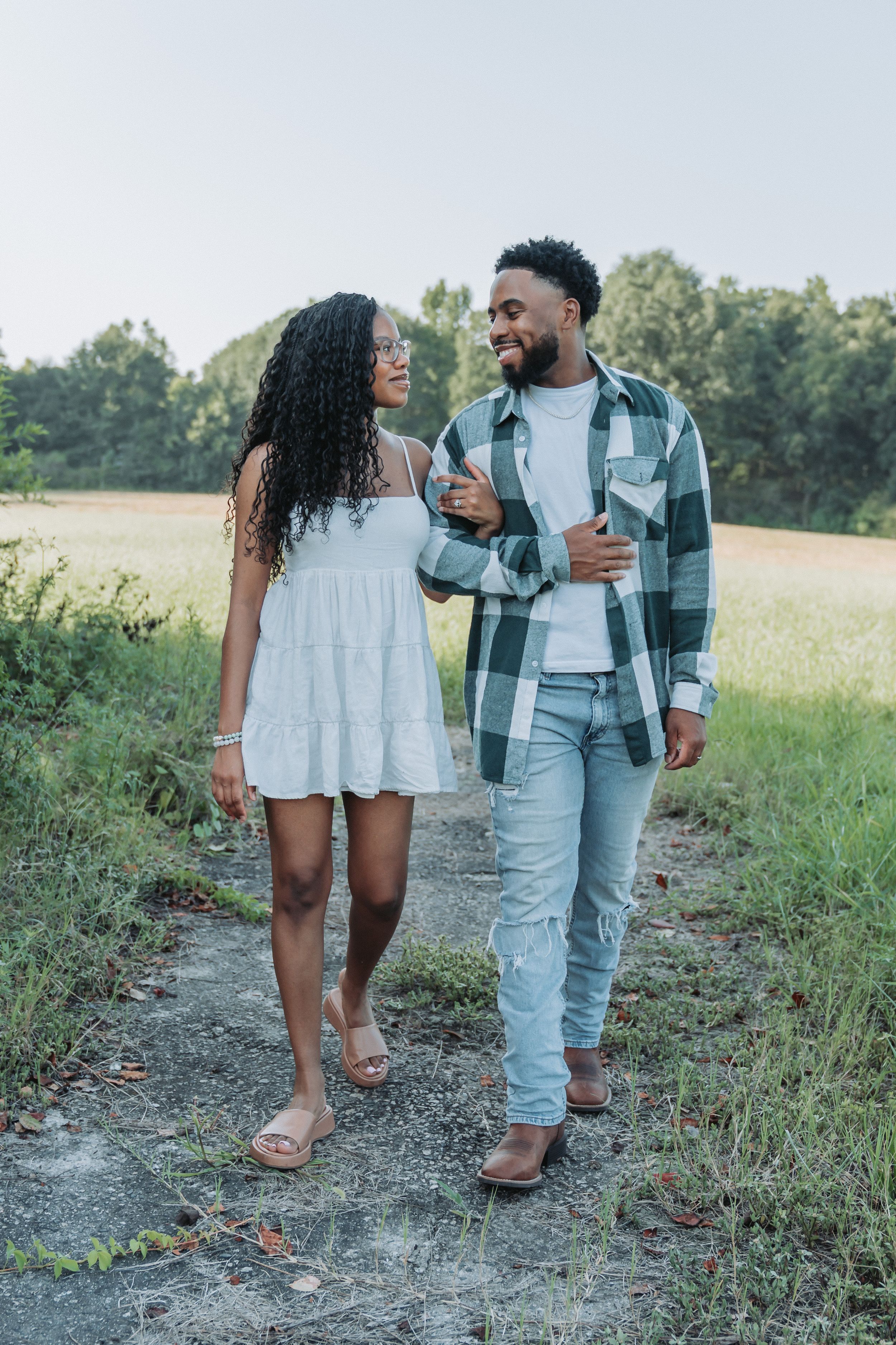 A happy Black couple walks arm in arm through a scenic field, she in a white dress, he in a plaid shirt and jeans.