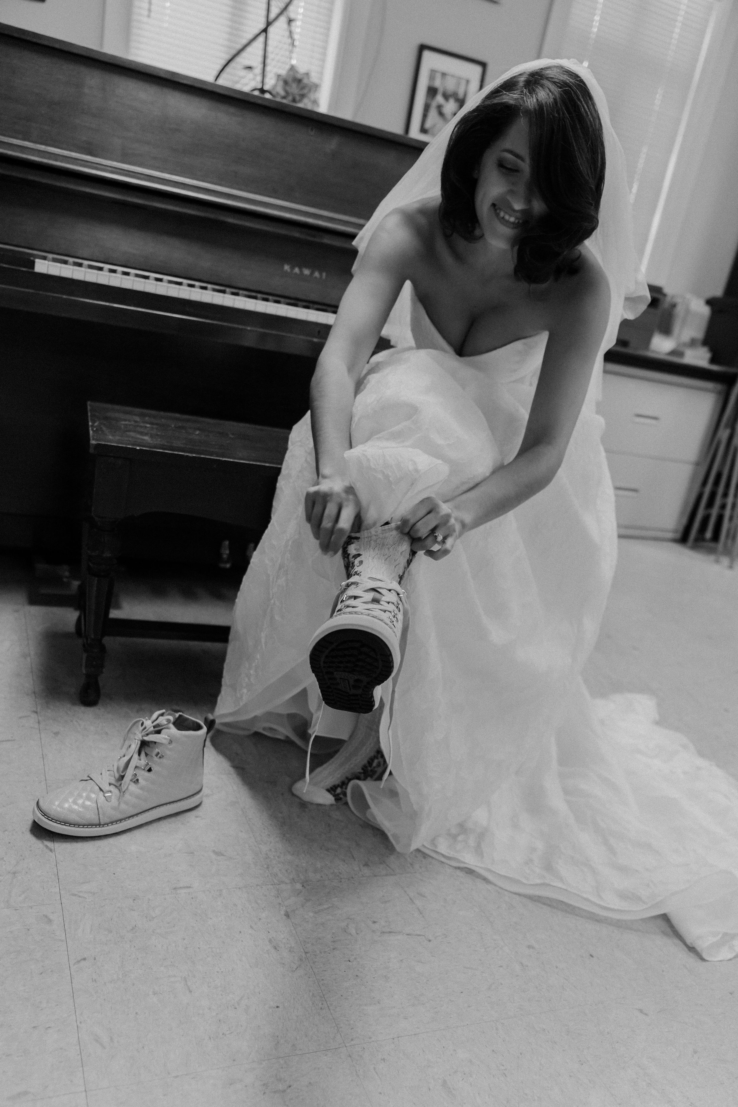 Bride in wedding gown smiling while putting on Converse sneakers beside a piano, black and white photo.