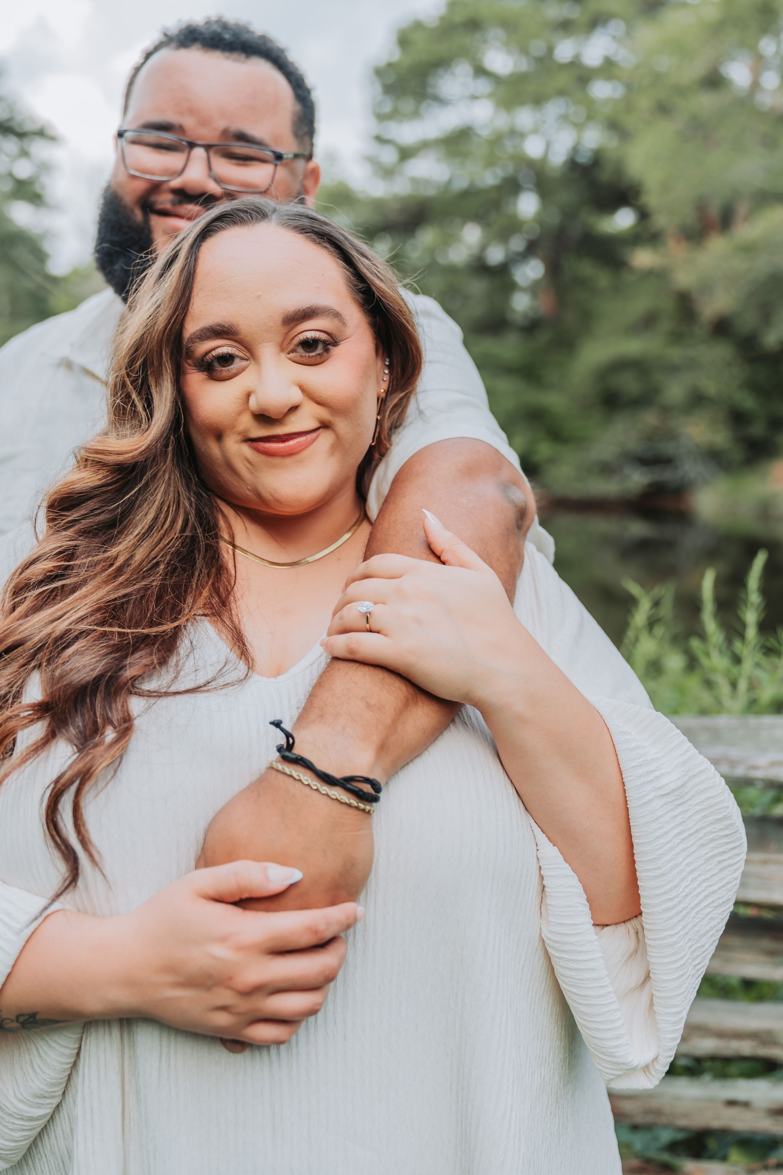 Couple embracing outdoors near a pond, woman showing engagement ring, wearing white outfits, smiling warmly.