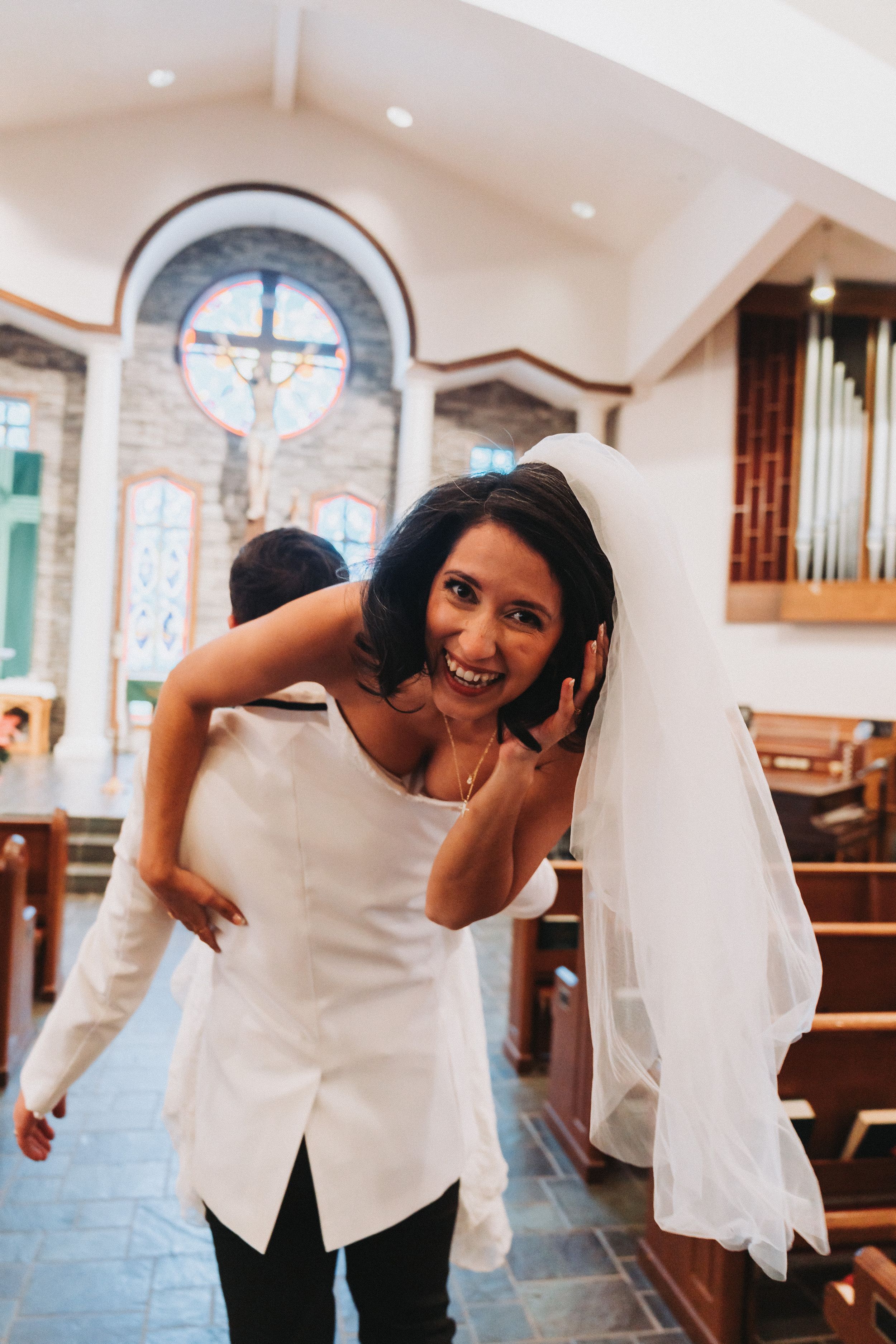 Smiling bride in white dress and veil being carried piggyback inside a church with stained glass windows.