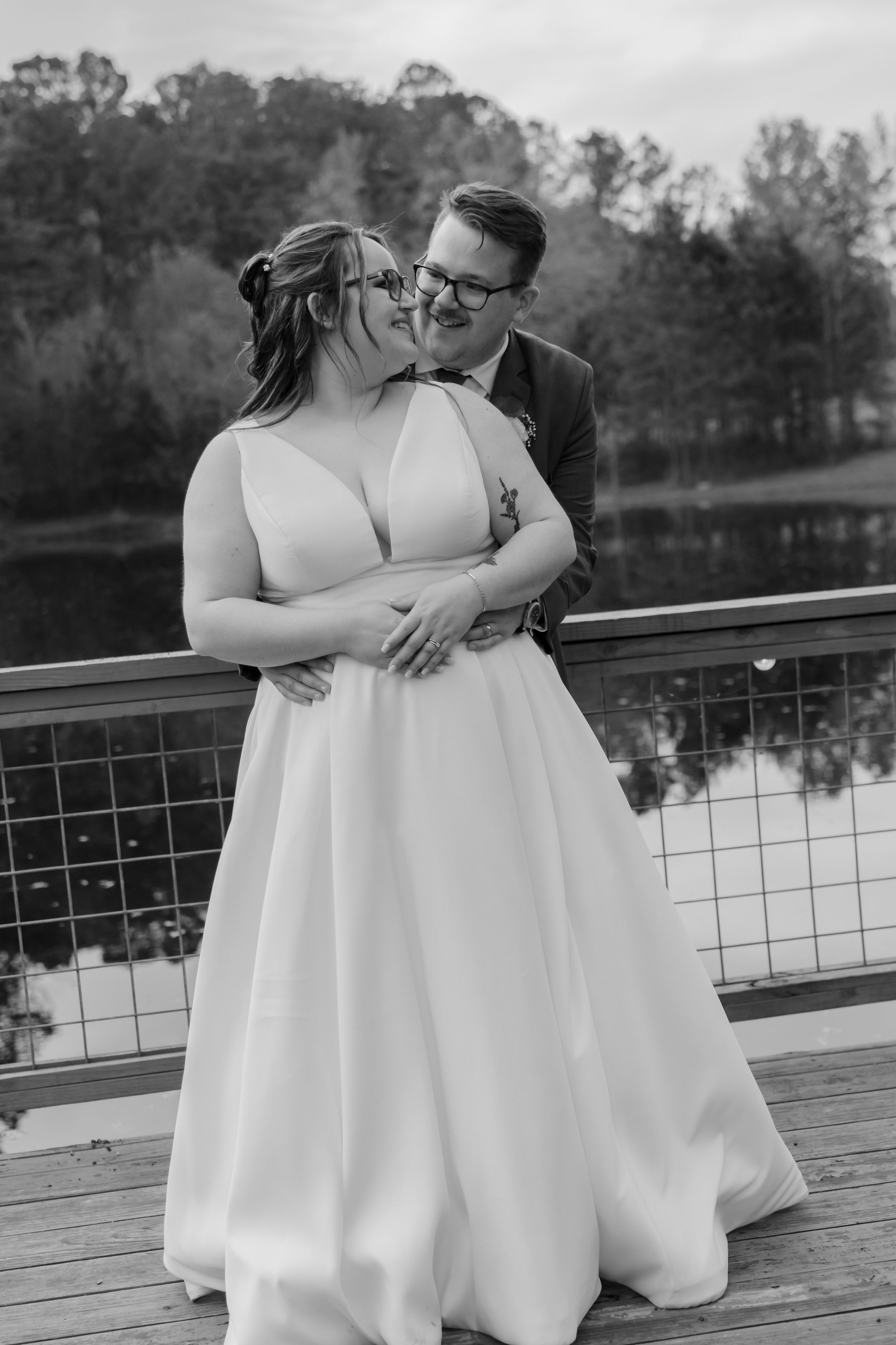 Black and white photo of a bride and groom sharing a tender kiss on a deck near a lake surrounded by trees.