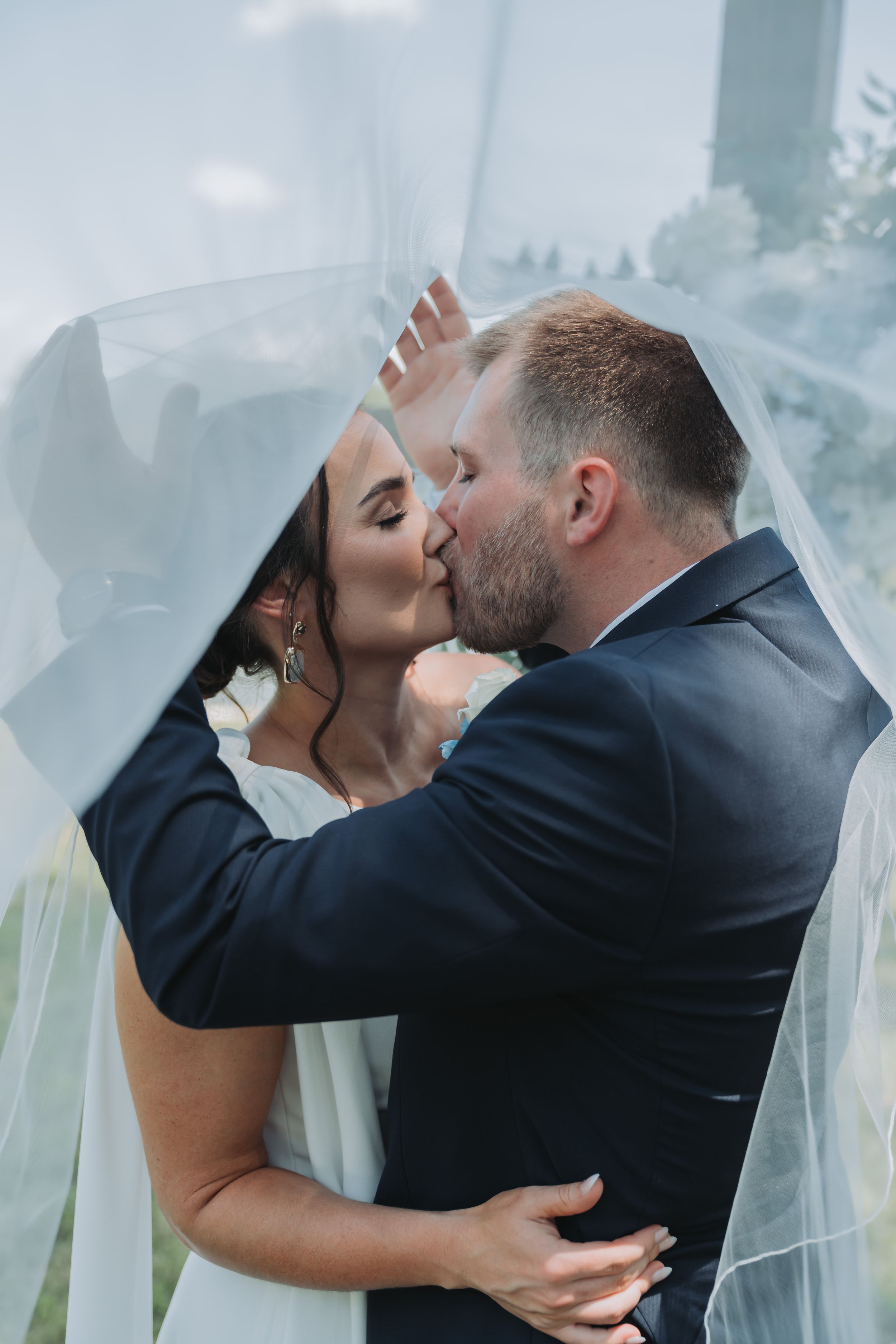 Bride and groom sharing a romantic kiss under a flowing wedding veil outdoors on their wedding day.