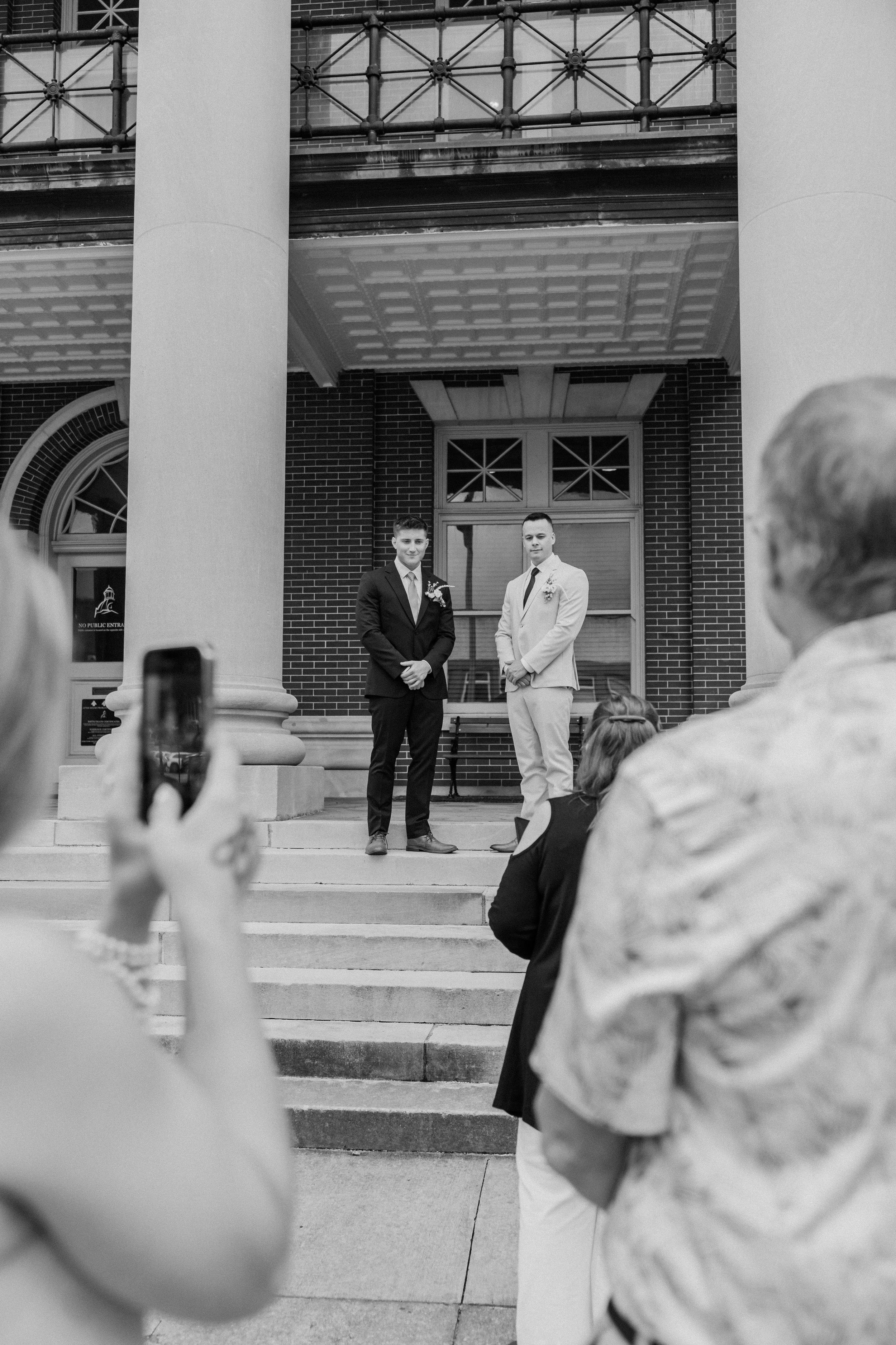 Black and white photo of two grooms posing on building steps as guests photograph their wedding ceremony.