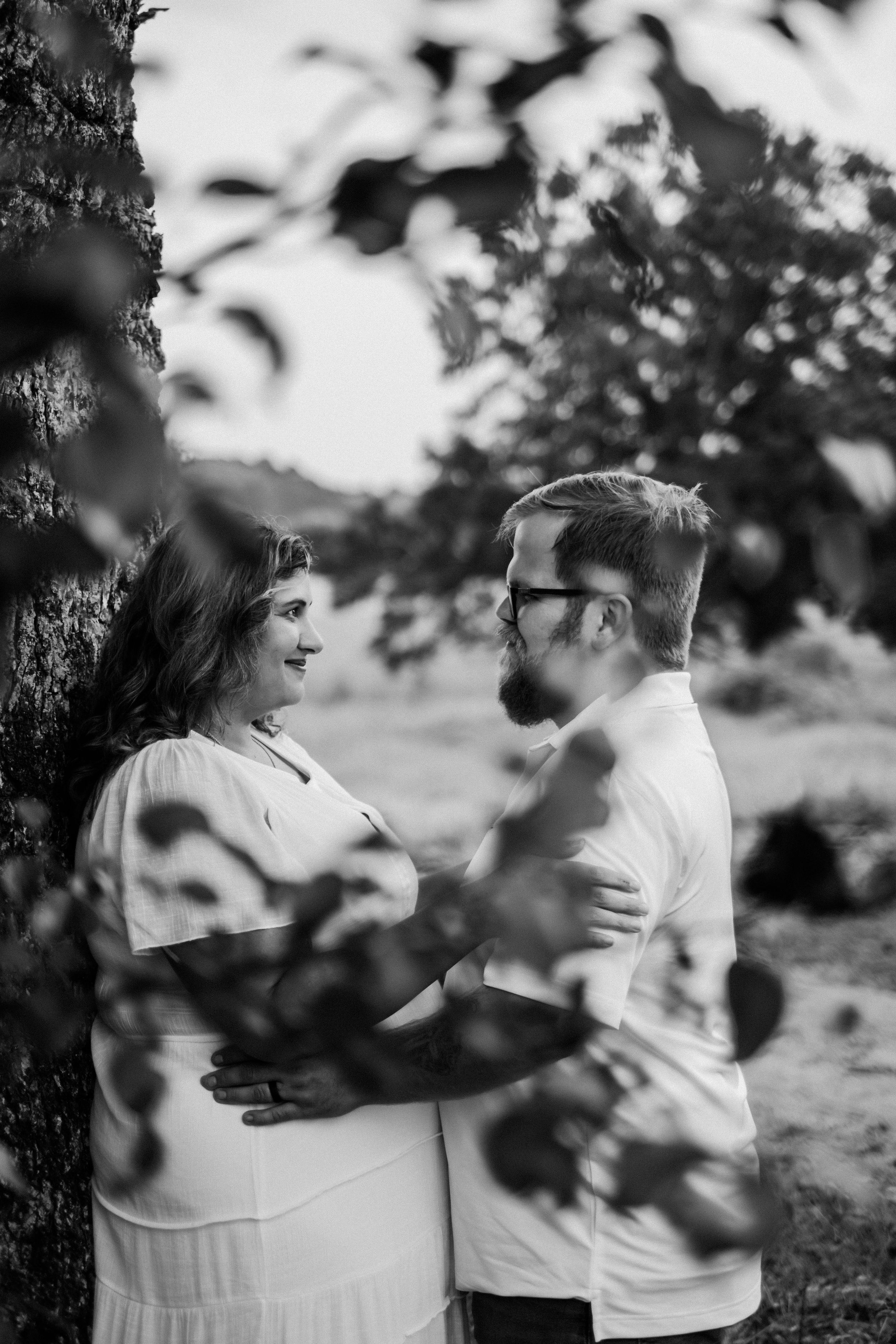 Black and white photo of a pregnant couple embracing near a tree outdoors, sharing a tender moment.
