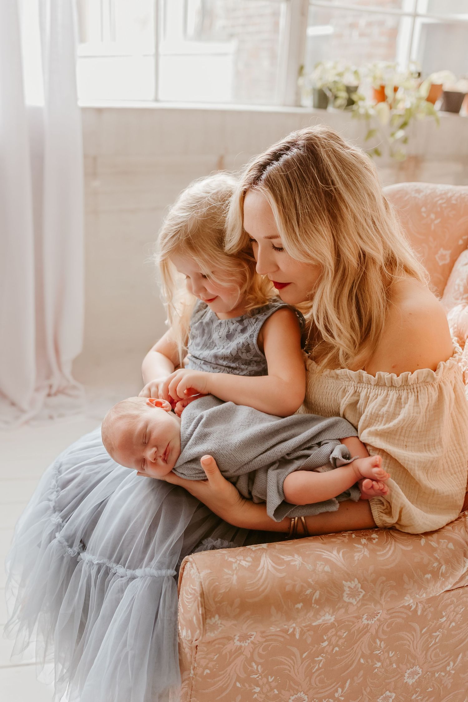 A mother with blonde hair sits in a floral chair holding a newborn while her young daughter in a gray dress leans in closely.